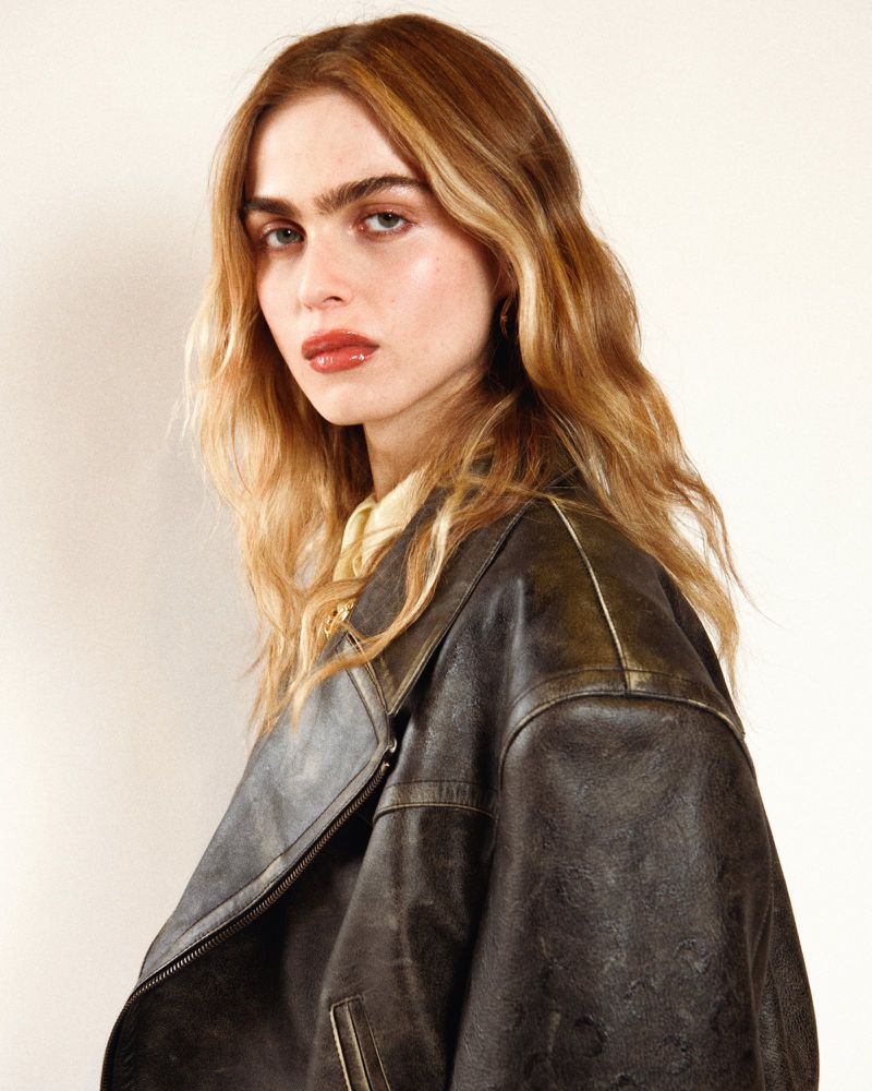 Adult female with fair skin poses indoors wearing dark leather jacket and yellow collared shirt against a neutral studio background.