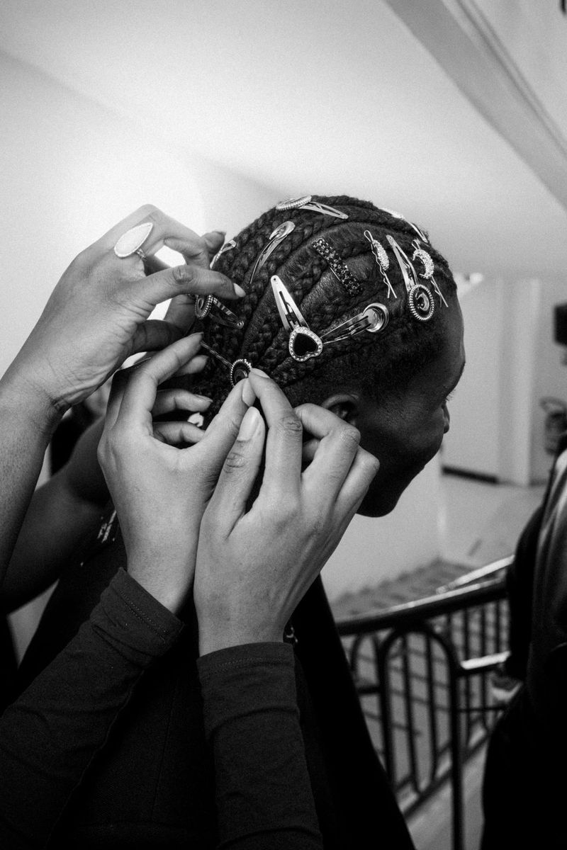 Black and white image captures an individual receiving hair styling with braids and metal clips, showcasing detail and texture.