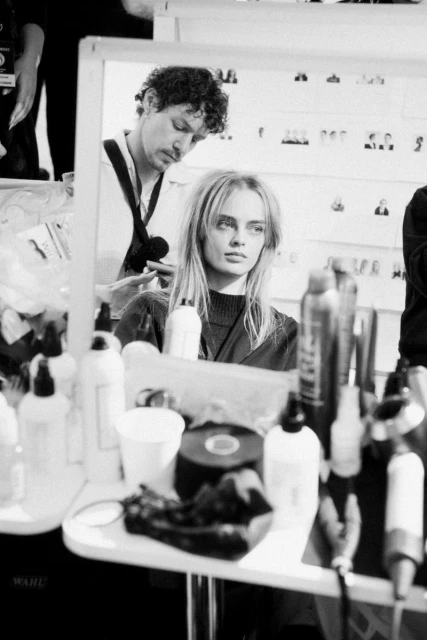 Backstage shot of a fashion model getting her hair styled while looking in a mirror at a fashion show, black and white photography.