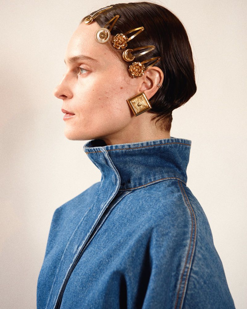 Profile of a female adult with golden hair clips and square earrings, wearing a denim jacket against a neutral background in a studio setting.