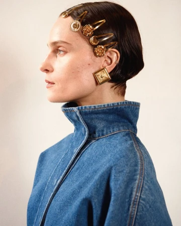 Profile of a female adult with golden hair clips and square earrings, wearing a denim jacket against a neutral background in a studio setting.