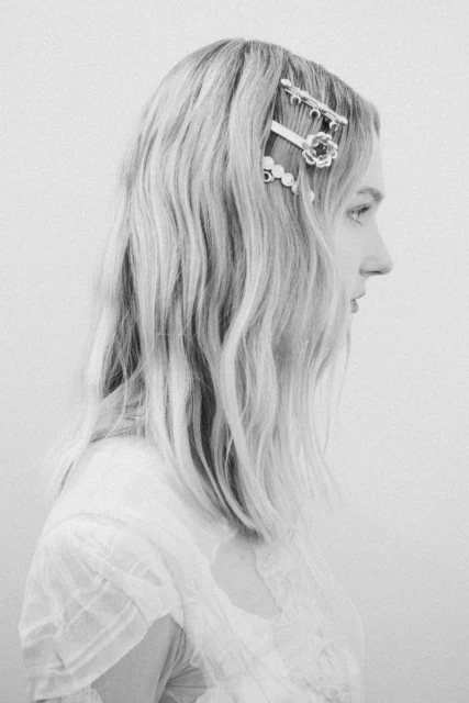 Monochrome portrait of a young woman in profile with blond wavy hair adorned with decorative hair clips and a light, sheer top.