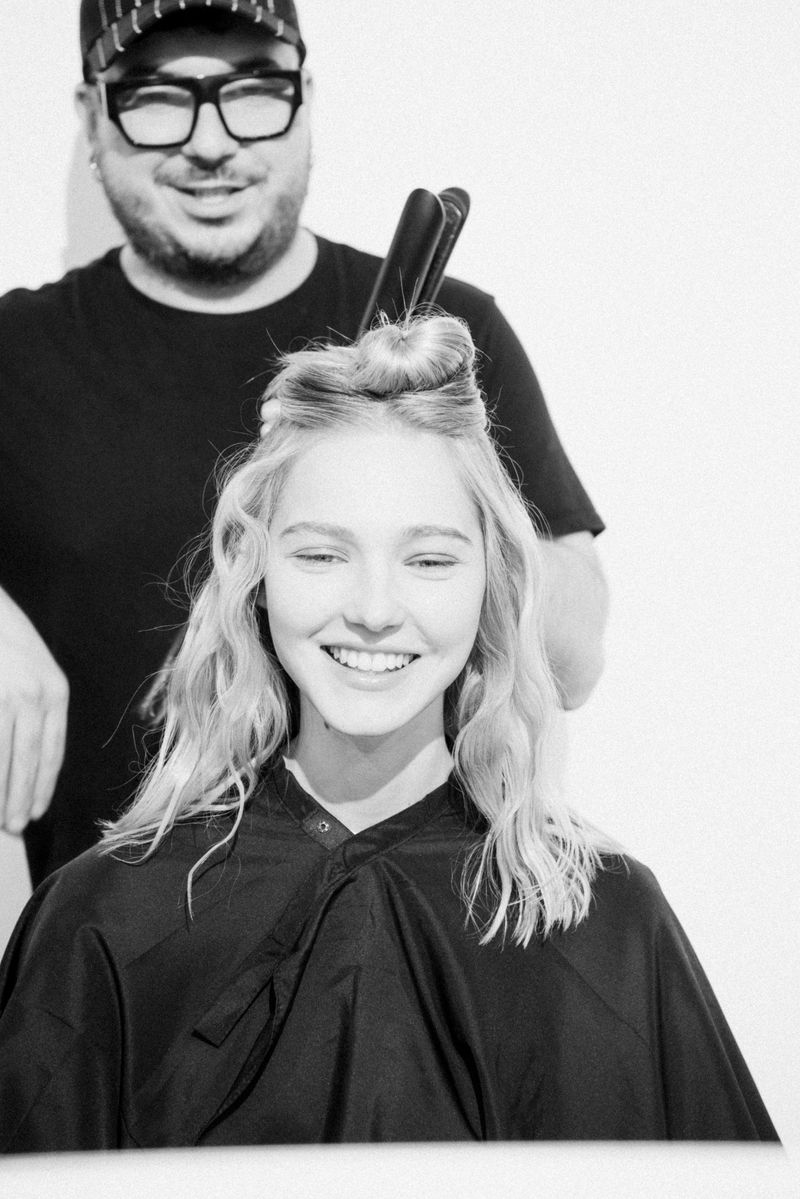 Black and white portrait of a hairstylist and a smiling young woman in a salon cape, getting her hair styled with a flat iron.