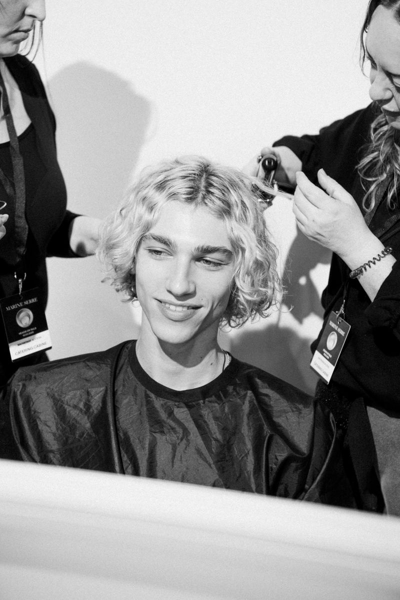 Backstage monochrome shot of a young, fair-skinned male model smiling as stylists work on his blonde, wavy hair for a fashion show.