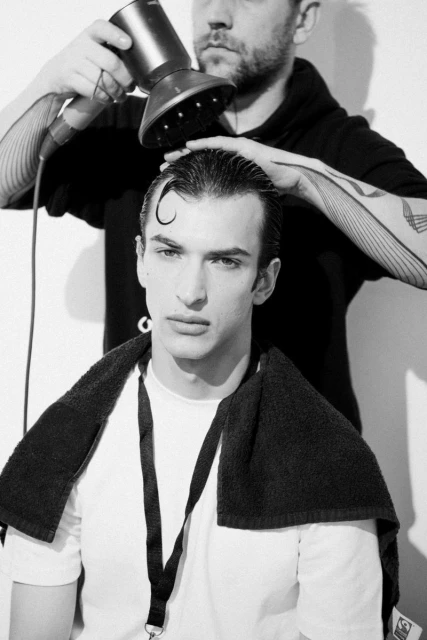 A stylist uses a diffuser on a young adult male with wet hair in a black and white fashion photo, set in a light studio.