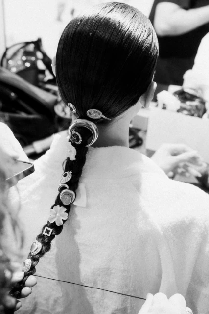 Back view of a young woman with a dark braided hairstyle adorned with decorative hair clips, wearing a white robe in an indoor setting.