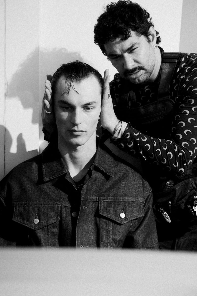 Black-and-white studio shot of a male model getting his hair styled by a second man with patterned clothing.