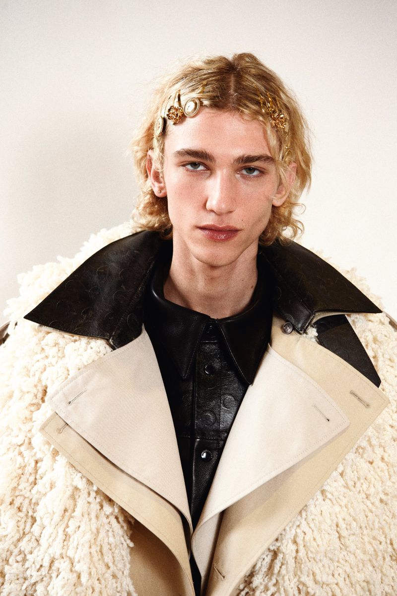 A single blonde adult male with fair skin poses indoors in an off-white sherpa coat with a black leather shirt and hair accessories.