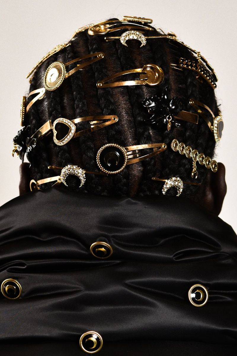 Close-up back view of a person with braids, gold and black hair clips, and a draped black garment with gold and black buttons.