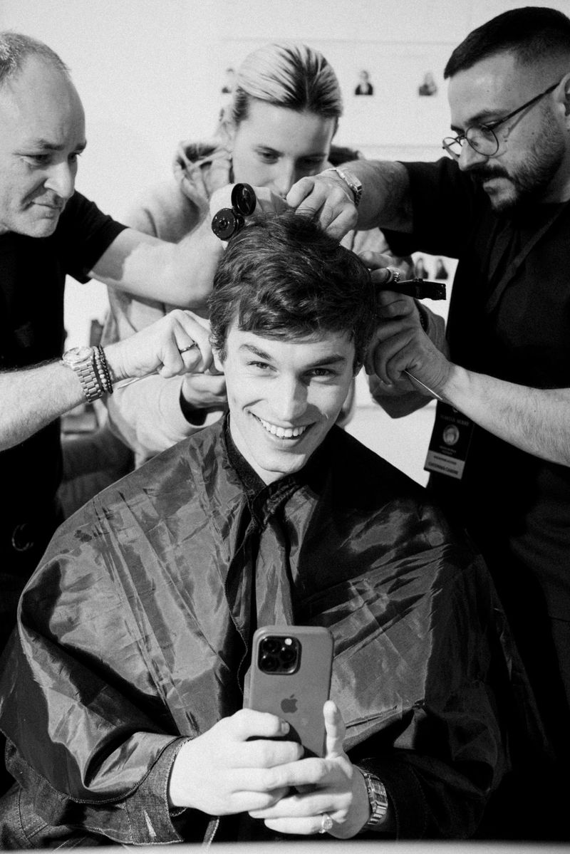 Smiling man takes a selfie in a barber cape, surrounded by three people styling his hair, in a black-and-white studio shot.
