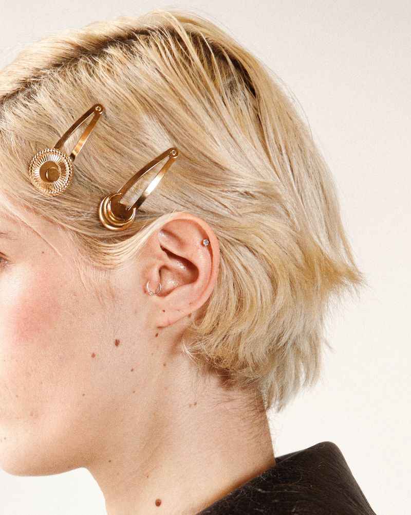 Close-up shot of a blonde woman's profile with golden hair clips. Beauty shot against an off-white background in a photo studio.
