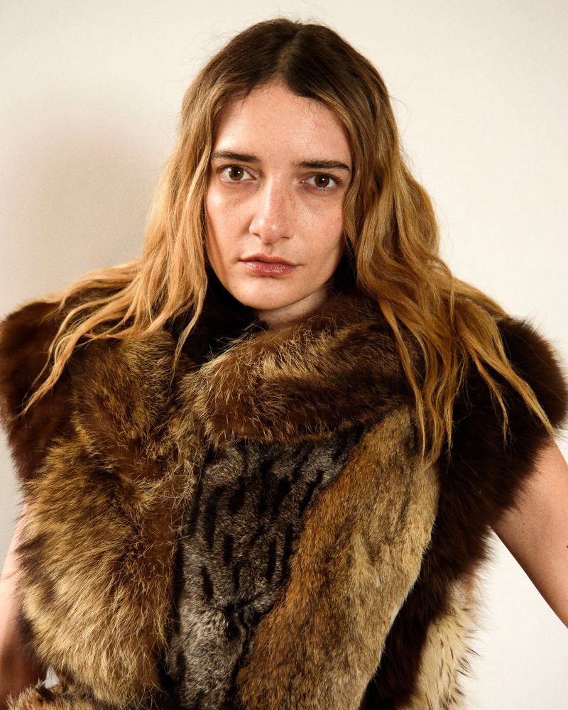 Portrait of a woman with long blonde hair wearing a fur vest against a plain white backdrop in a studio setting.