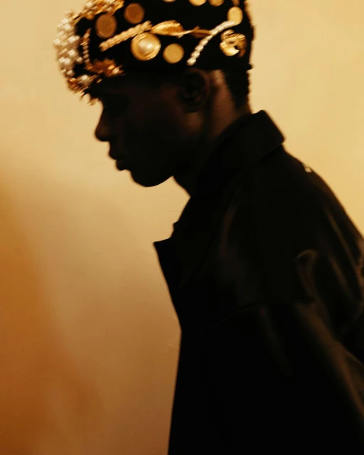 Close-up of a Black male model in profile wearing a decorative beaded and gold-coin-studded headdress, shot in warm, diffused lighting.