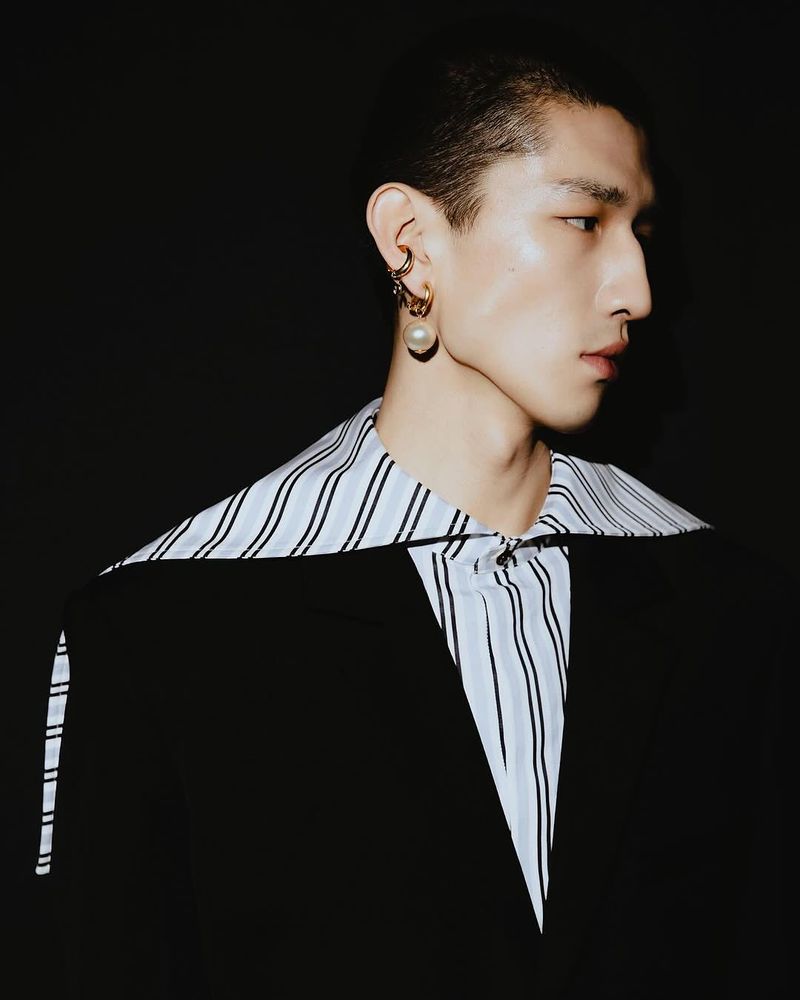 Portrait of an Asian man with short dark hair wearing a black blazer and striped shirt against a dark background in a photo studio.