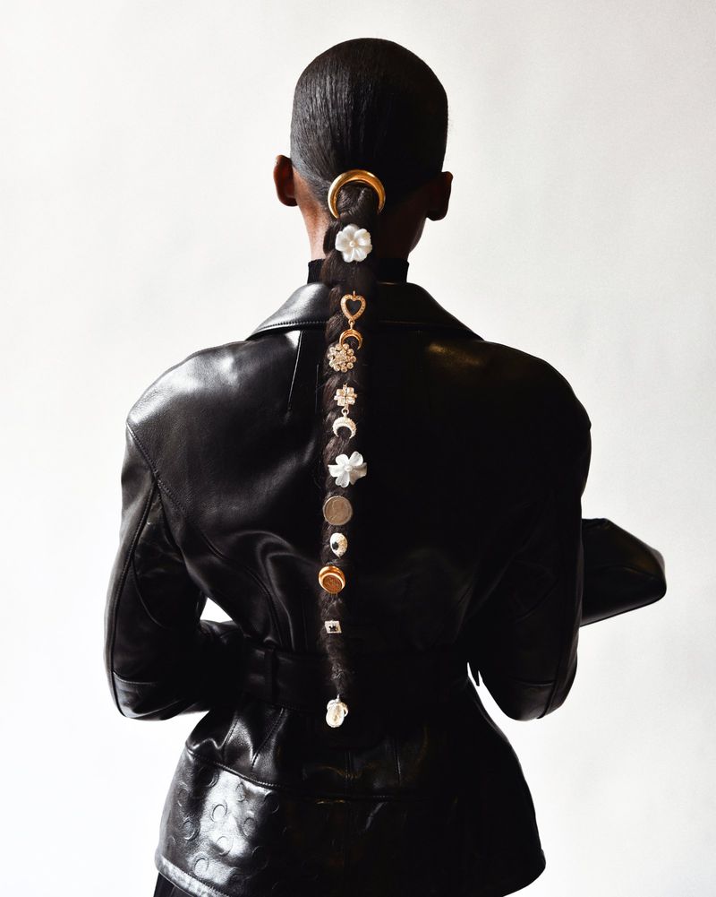 Back view of Black model in a black leather jacket with a braided hairstyle adorned with flower and coin charms against a white background.