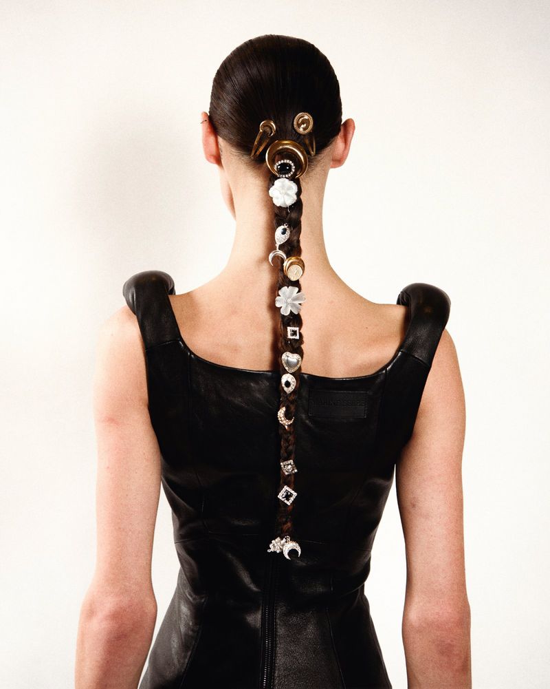 Back view of a slender woman in a black dress with a braided hairstyle adorned with diverse jewelry against a plain white backdrop.