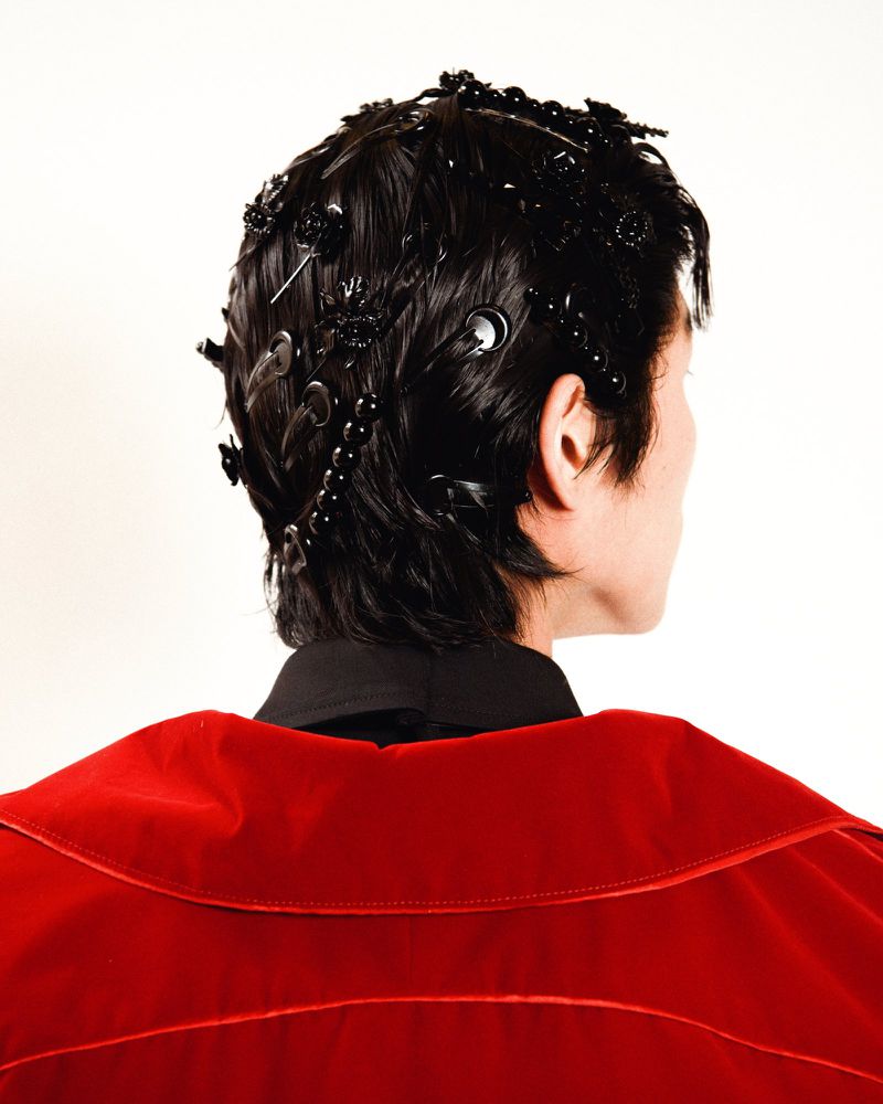 An adult with black hair and accessories poses in front of a white background, wearing a red velvet jacket and a black collared shirt.