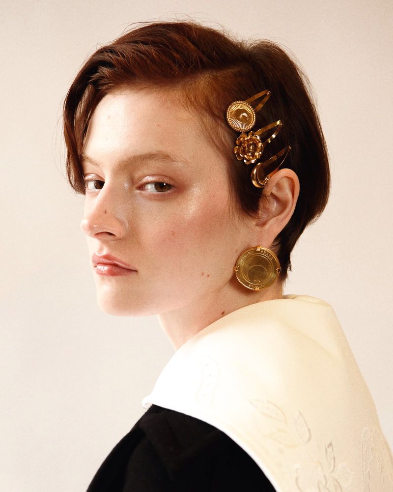 Portrait of a young woman with auburn hair styled with gold barrettes, wearing large disc earrings, and a white and black dress against a white backdrop.