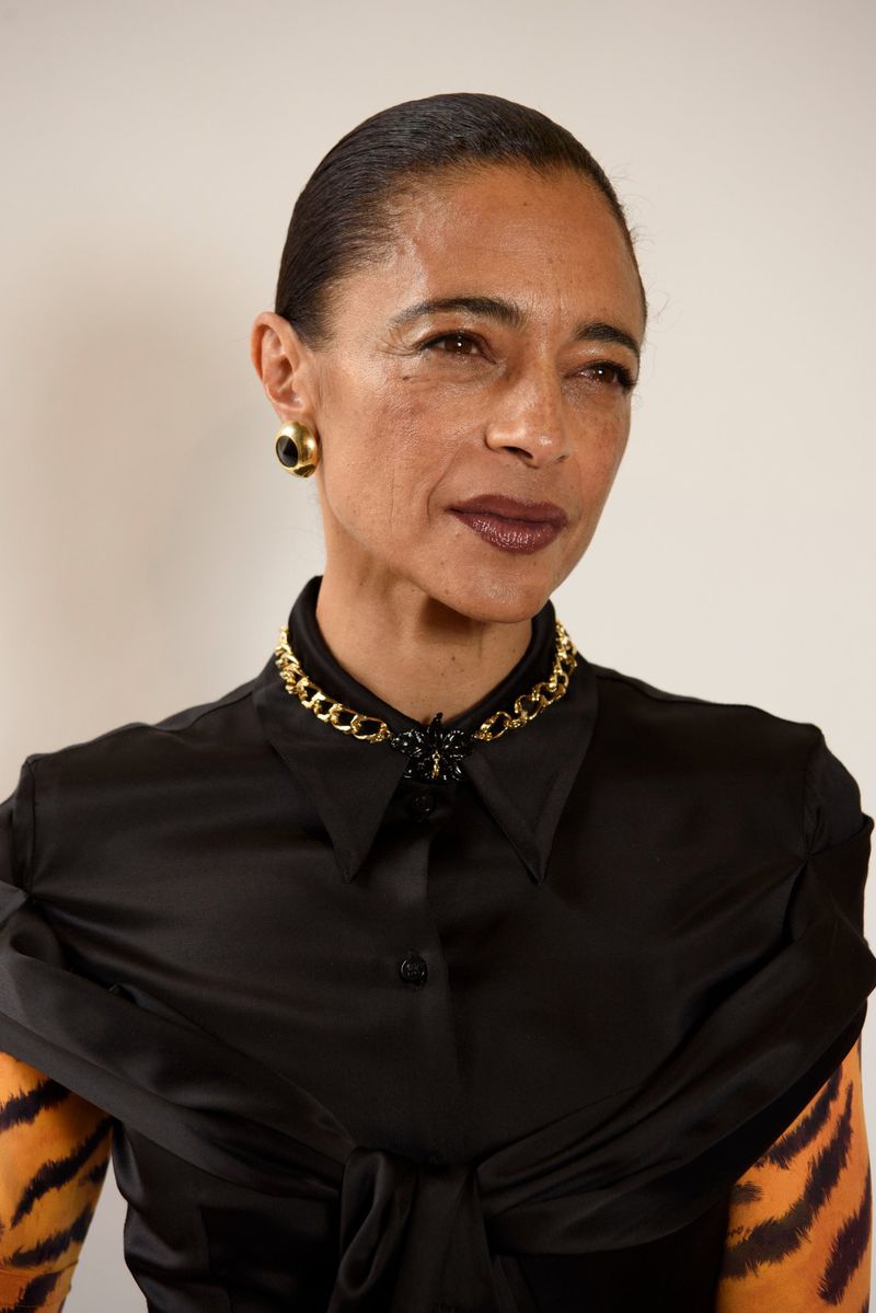 Close-up portrait of an adult woman with dark hair and skin wearing a black satin blouse and gold jewelry against a plain background.