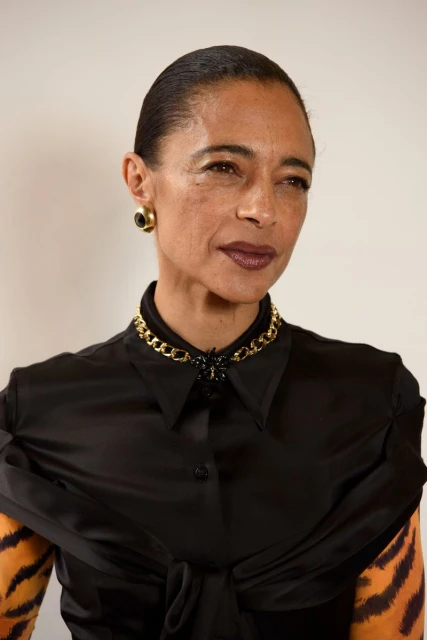 Close-up portrait of an adult woman with dark hair and skin wearing a black satin blouse and gold jewelry against a plain background.