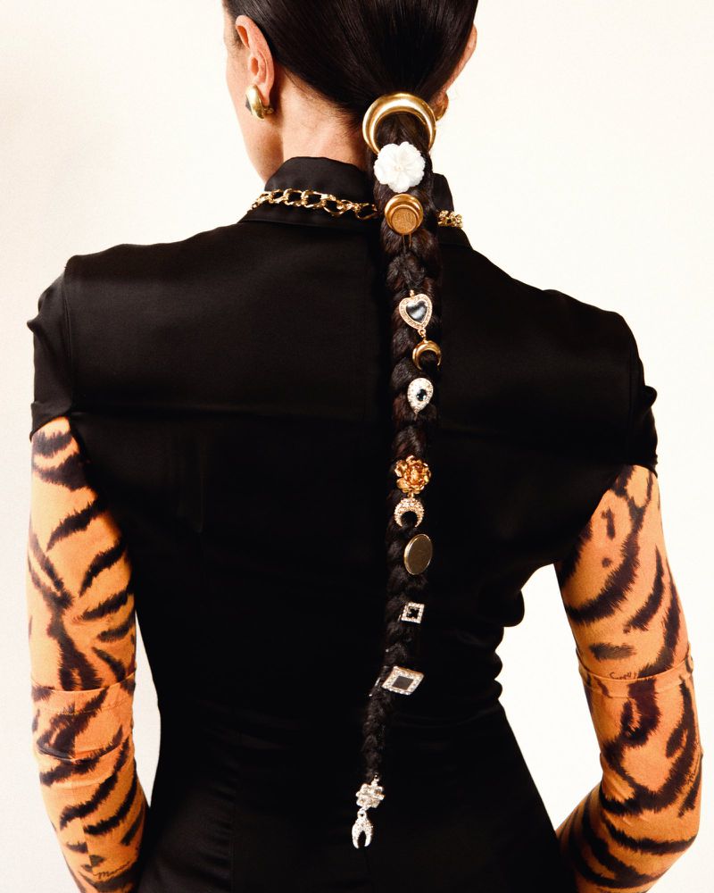 Back view of a woman with a long, dark braid adorned with gold and silver charms and accessories, wearing a black dress and tiger-print sleeves.