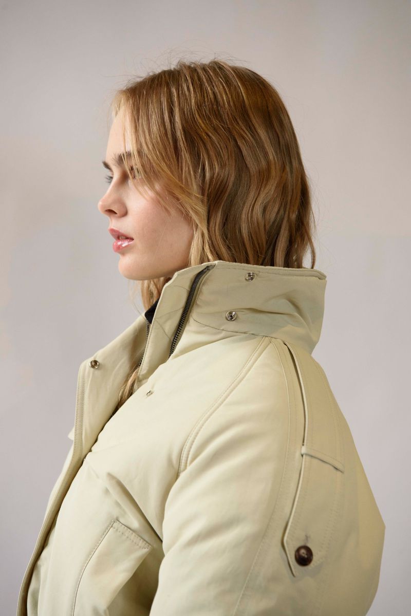 Portrait of a young, light-skinned woman in a profile view, showcasing wavy blonde hair and wearing a light beige puffer jacket indoors.