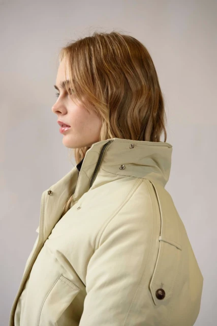 Portrait of a young, light-skinned woman in a profile view, showcasing wavy blonde hair and wearing a light beige puffer jacket indoors.