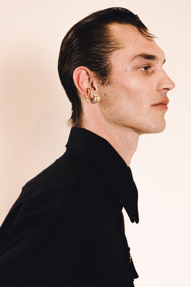Adult man in a black collared shirt and gold earrings poses in a studio with a side profile. He has dark hair slicked back.