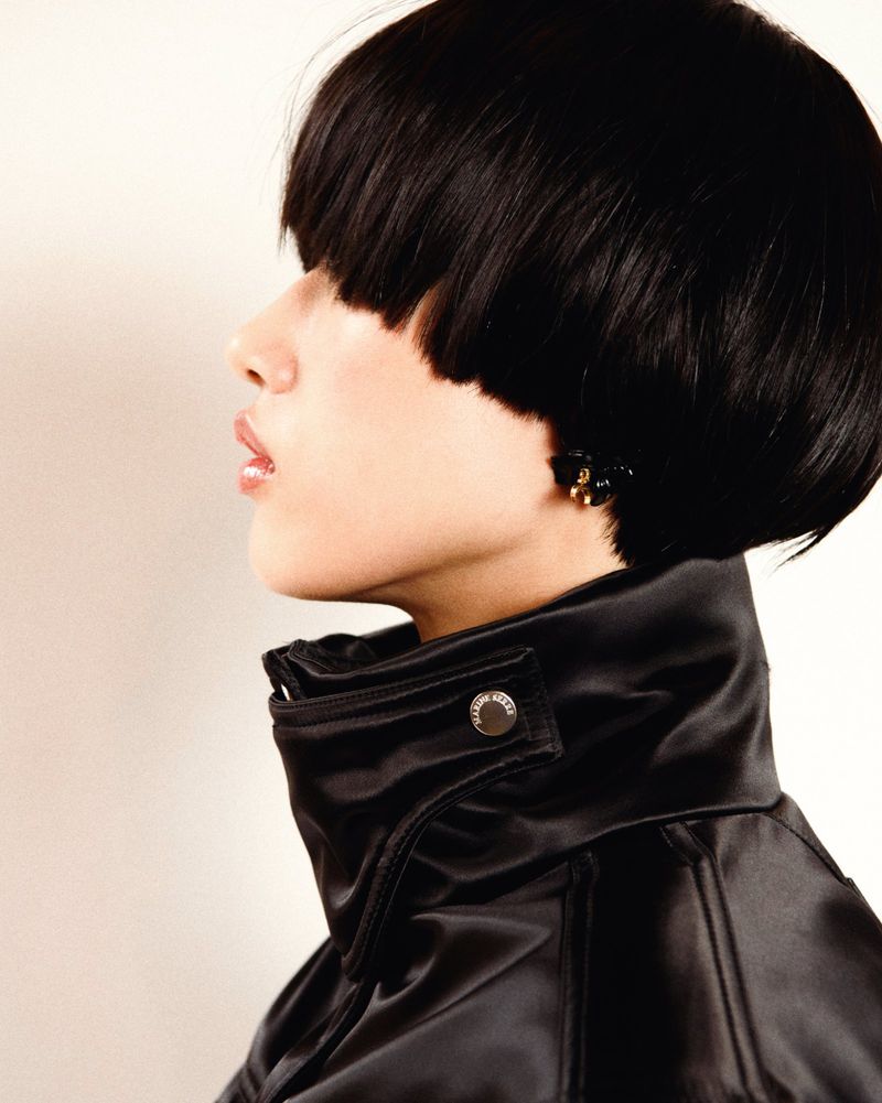 Woman in black satin jacket with a short black bob hairstyle posing in profile against a pale beige backdrop in a fashion photography.
