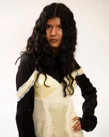 Model with long dark wavy hair wearing a pale yellow lace slip dress over a black long-sleeve top against a white background