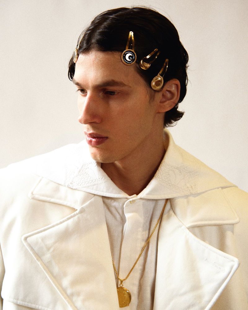 Portrait of a male fashion model in a white jacket, adorned with golden hair clips and a matching necklace, standing against a plain background.