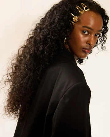 Model with long dark curly hair wearing gold hairpins and a black jacket against a plain background