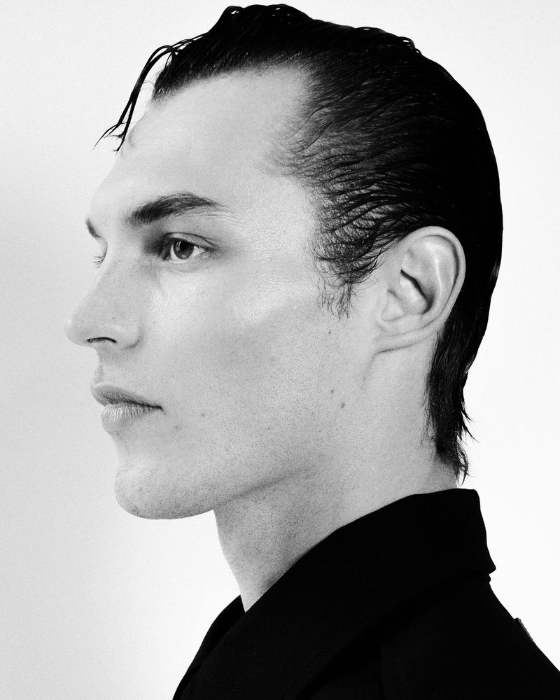 Striking black and white portrait of a young adult male with slicked-back dark hair in profile against a clean, bright white backdrop.