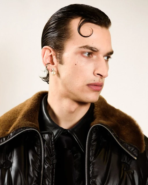 Close-up of a stylish adult male model with slicked-back hair wearing a black jacket with a fur collar and subtle pearl earrings.