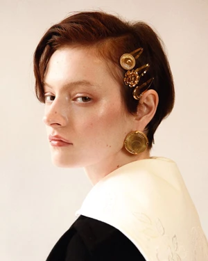 Close up portrait of a short haired model wearing gold hair clips and round gold earrings against a light background