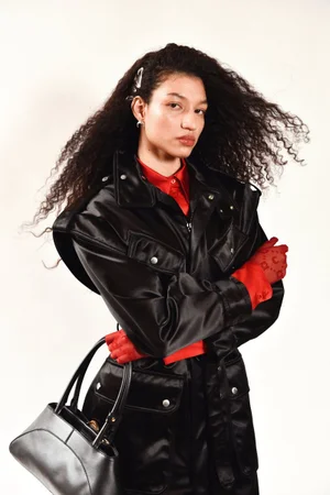 Model with long dark curly hair wearing a black leather coat holding a black handbag against a white background