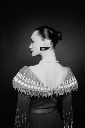 Model in profile wearing an embellished dress with a beaded collar, pearl necklace, and large earring against a dark background