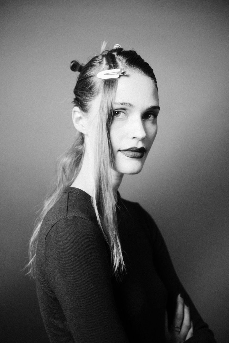 Black and white portrait of a woman with hair clips, wearing a dark top, looking directly at the camera with arms crossed.