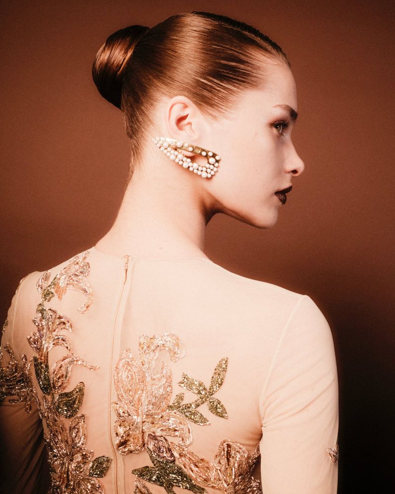 Woman in a beige dress with embroidered flowers and a pearl hair clip poses in a studio with a warm, brown background.