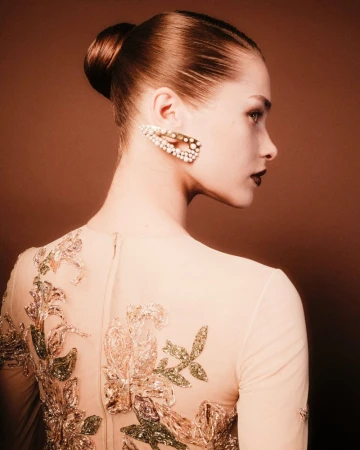 Woman in a beige dress with embroidered flowers and a pearl hair clip poses in a studio with a warm, brown background.