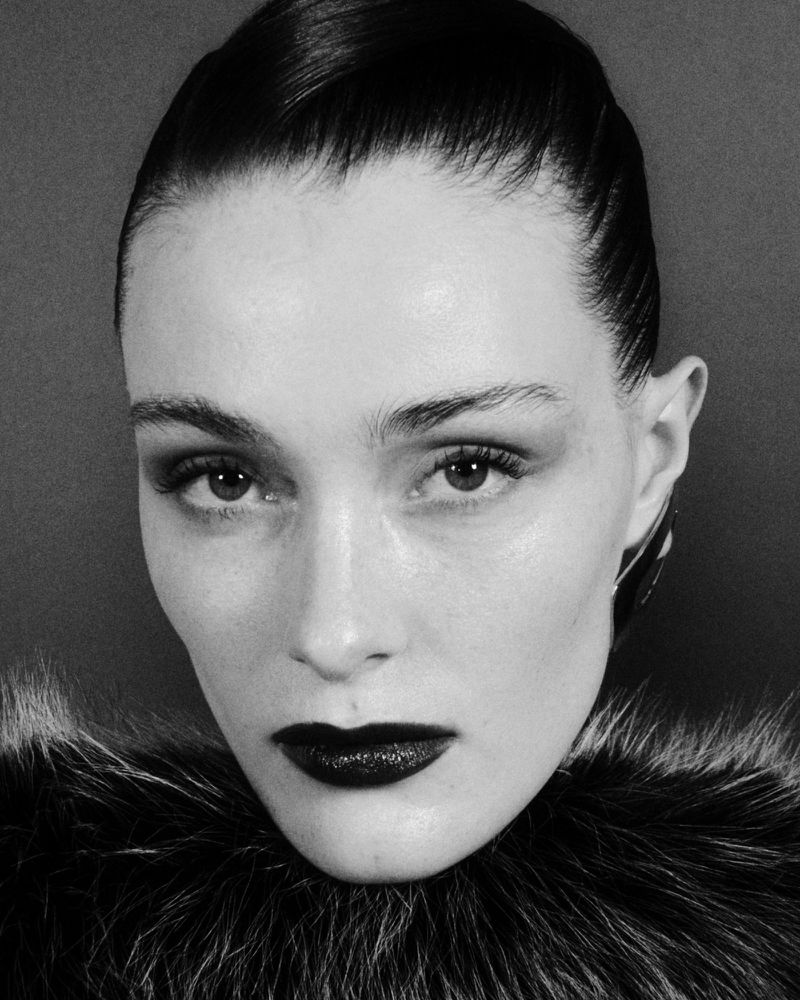 Monochrome close-up portrait of a woman with sleek dark hair, dramatic makeup, and a dark fur collar, against a dark grey background.