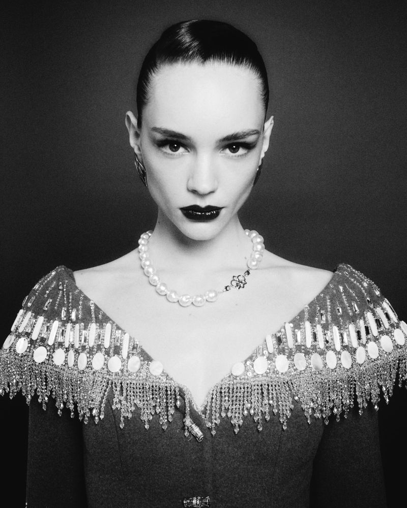 Black and white portrait of a woman with dark hair, wearing a pearl necklace and an embellished dress with crystal fringe details.