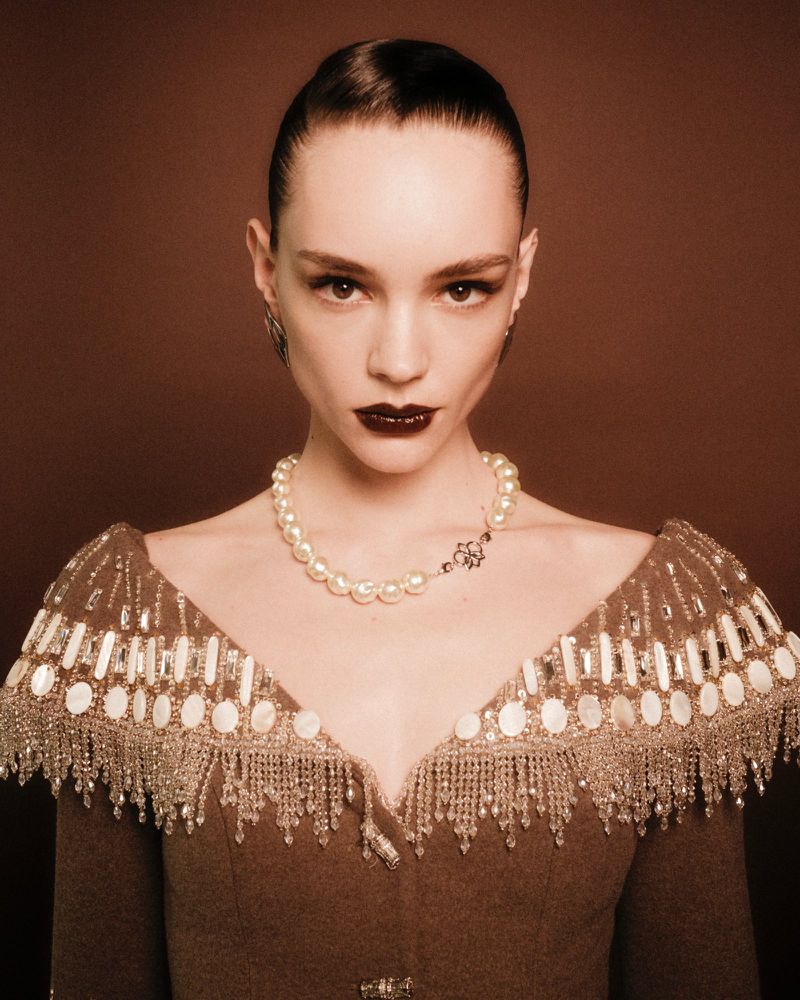 Portrait of a female fashion model with dark lipstick and elegant, beaded pearl-embellished couture against a muted brown backdrop.