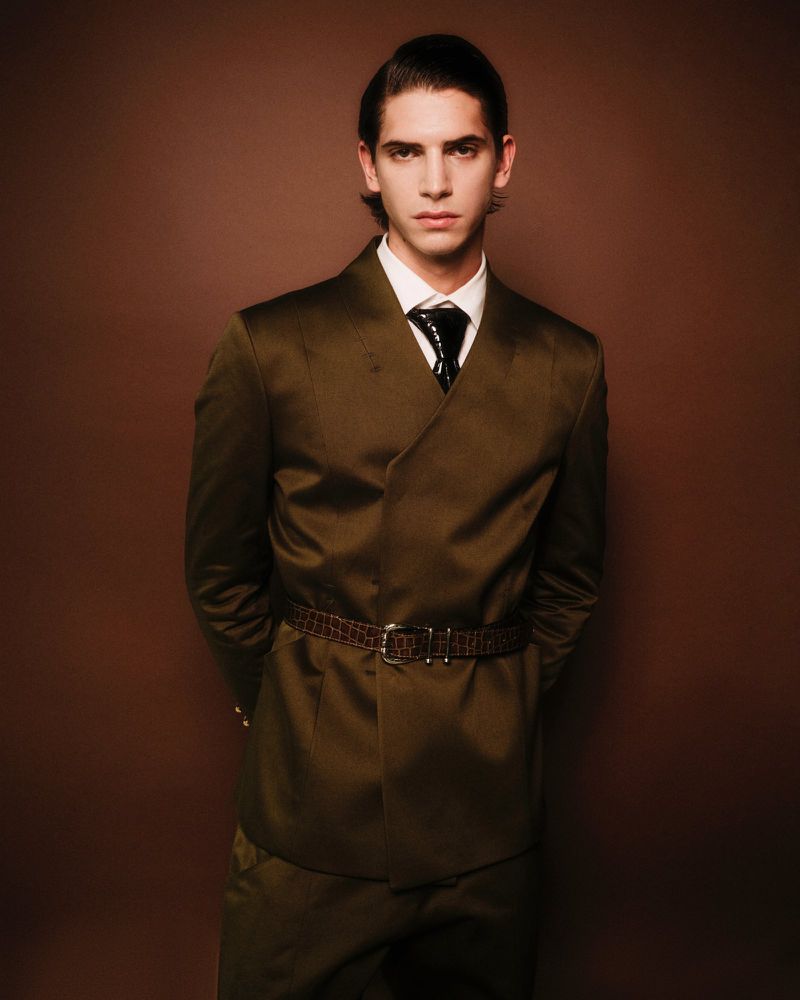 Portrait of a handsome male fashion model in a stylish olive-green suit with a thin brown belt, posing against a brown background.
