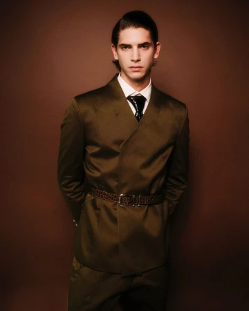 Portrait of a handsome male fashion model in a stylish olive-green suit with a thin brown belt, posing against a brown background.