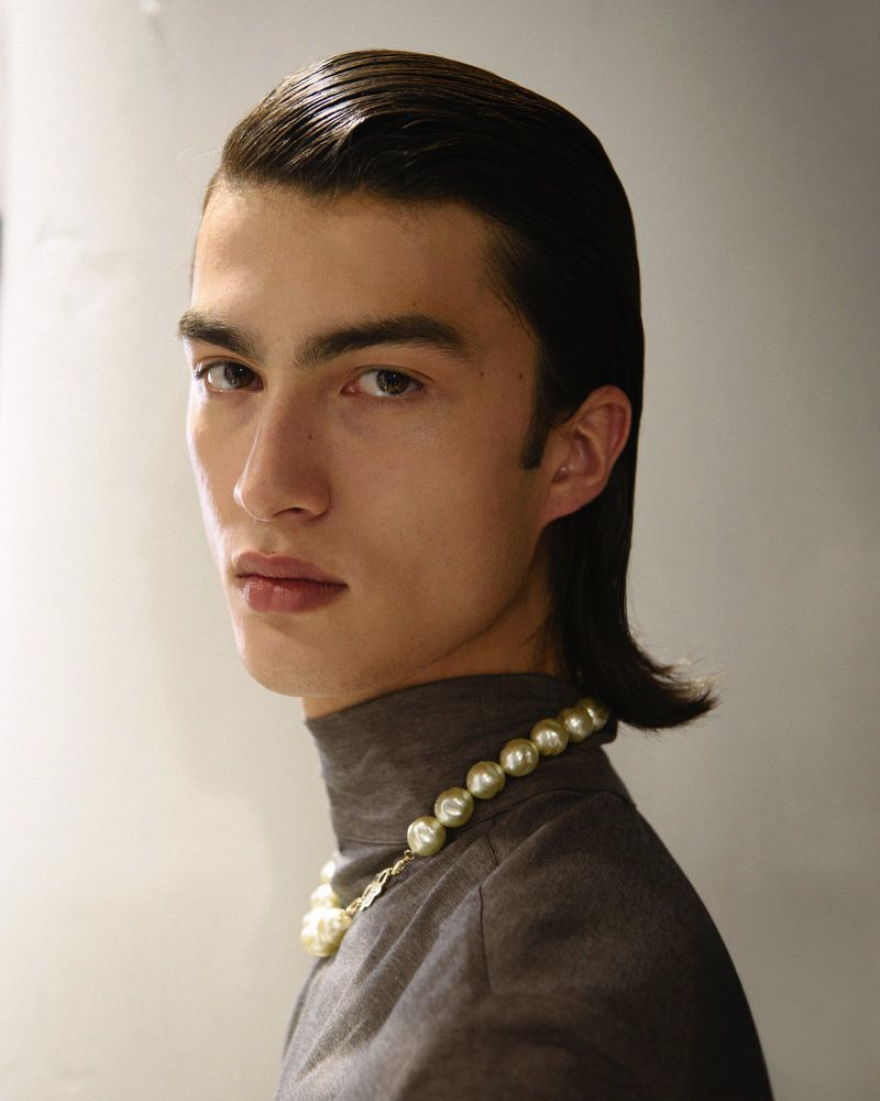 Close-up portrait of a young adult male with dark hair, wearing a grey turtleneck and a large pearl necklace, against a neutral wall.
