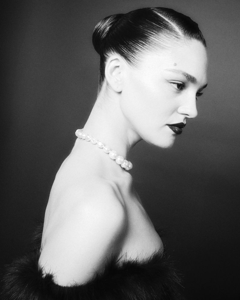 Monochrome portrait of a woman with slicked-back hair, pearl necklace, and a feathered shawl against a plain dark backdrop.