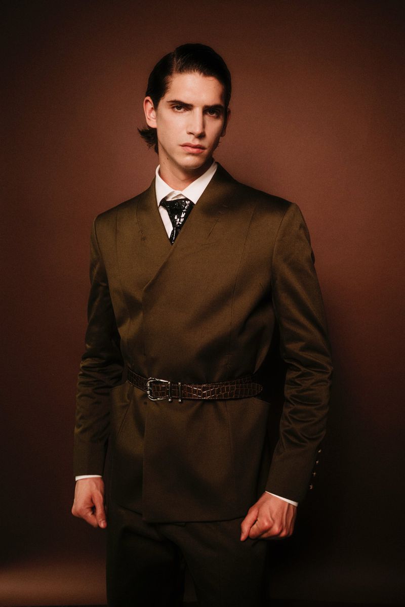 Male fashion model in an olive green suit with a belt, white shirt, and patterned tie poses in a studio with a warm brown backdrop.