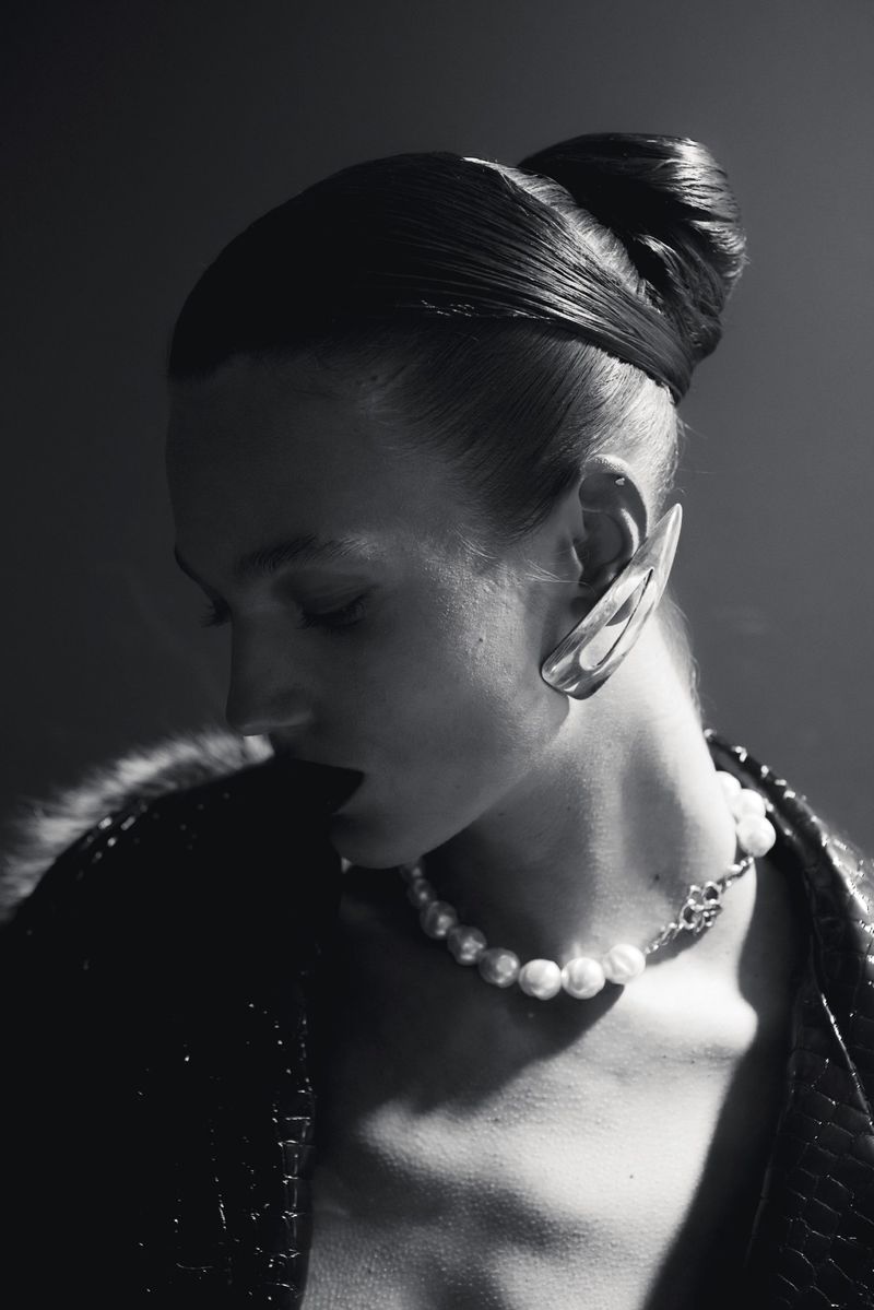 Monochrome portrait of a woman with styled hair, large ornamental ear cuff, pearl necklace, and sequined jacket against a dark backdrop.