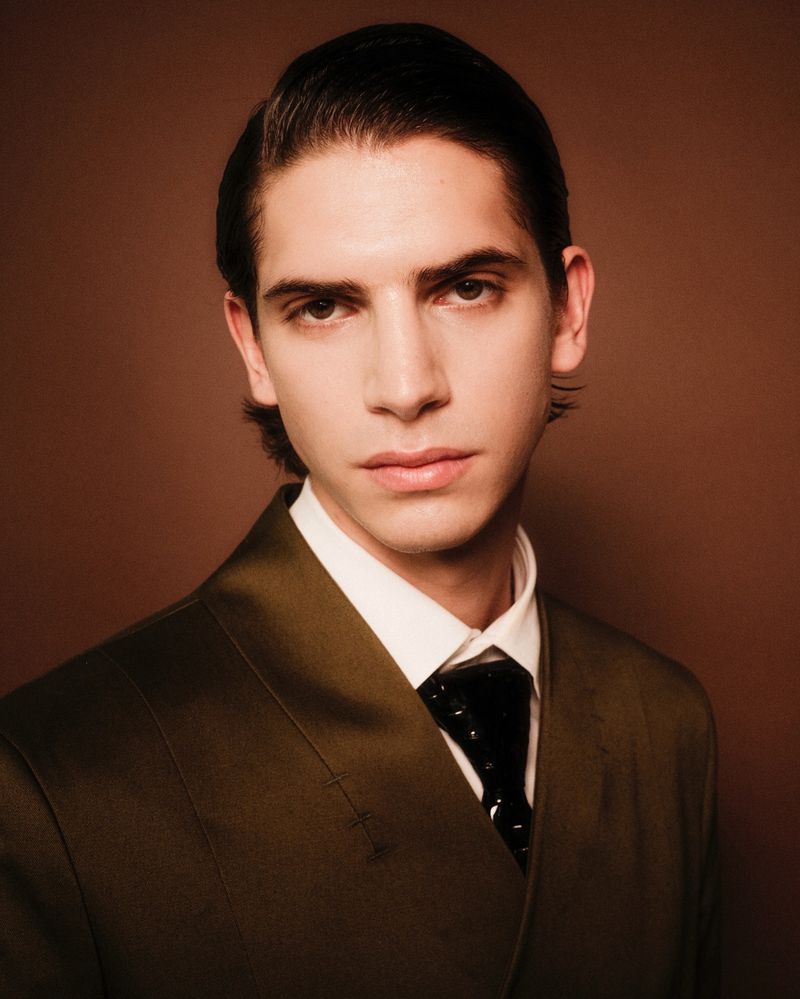Headshot of a stylish adult male with dark hair, fair skin, and a serious expression, wearing a suit and tie against a brown backdrop.